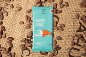 A teal chocolate bar wrapper, picturing an illustrated depiction of the country Dominican Republic, sits on a burlap sack among scattered cocoa beans.